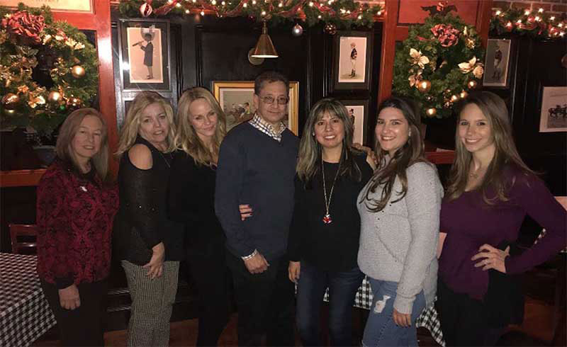 The image shows a group of people posing together for a photograph in front of a festive holiday setting, with a Christmas tree and decorations visible in the background.