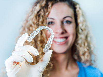 The image features a person holding up a transparent plastic object that appears to be a dental retainer or aligner, with the individual smiling and wearing a white lab coat.
