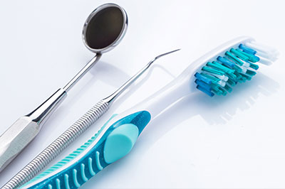 The image shows a collection of dental tools, including a toothbrush with blue bristles, placed next to a pair of metal tweezers and a small spoon-like instrument, all set against a white background.