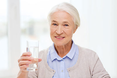 The image features an elderly woman holding a glass of water, smiling gently at the camera.