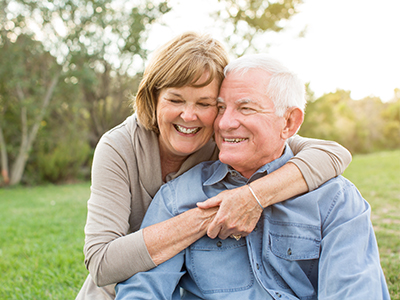 The image depicts an elderly couple embracing, with the man standing behind the woman. They are outdoors during daylight hours, and both individuals appear to be smiling.