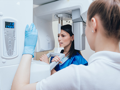 A woman in a blue lab coat stands next to a large, modern 3D scanner machine, with a technician overseeing the setup.