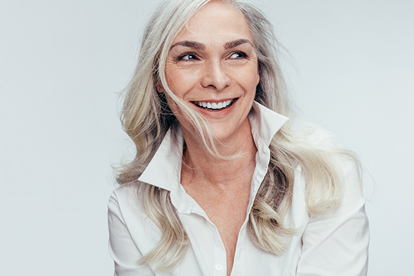 The image is a portrait of an older woman with blonde hair, wearing a white top and smiling.