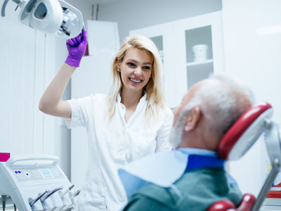 A dental hygienist is assisting an elderly man in a dental chair, with the woman holding a dental mirror and smiling.