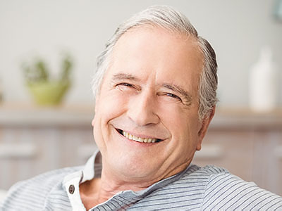 An elderly man with white hair is smiling and looking directly at the camera, wearing a blue shirt and seated in an indoor setting.